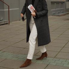 A woman wears a long gray coat, white straight-leg jeans, and tan suede ankle boots.