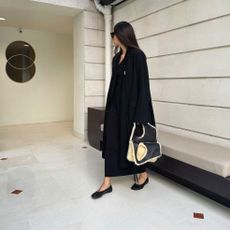 A woman wears a long black coat, black ballet flats, and a leather and shearing bag.