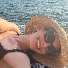 Anne Hathaway laying on a towel at the beach wearing a straw bucket hat and large sunglasses.