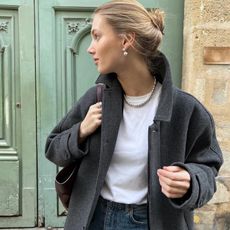 female Scandinavian model Moosgaard Nielsen poses in front of an ornate light green door with a messy bun, Lie Studio drop earrings, Lie Studio ball necklace, a white tee, gray coat, and jeans