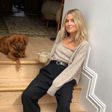 British influencer Lucy Williams poses on steps in her London home with her dog holding a cup of tea wearing a neutral cardigan and matching top, black belt, and black pants