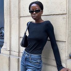 Influencer wearing black long sleeve top, black sunglasses, and straight-leg jeans while leaning up against Paris wall.