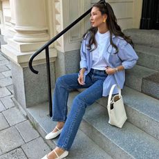 female fashion influencer Felicia Akerstrom poses on steps outside a building in Stockholm, Sweden wearing yellow gold earrings, a white t-shirt, blue striped button-down shirt, white mini tote bag, straight-leg jeans, and white Mary Jane ballet flats