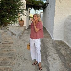 Fashion influencer Lucy Williams on summer vacation in Europe wears drawstring white pants, a breezy short-sleeve blouse, and flat strappy sandals.