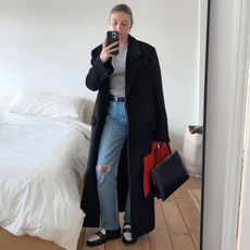 British fashion influencer Brittany Bathgate poses for a mirror selfie wearing a black maxi coat, gray t-shirt, black belt, ripped distressed jeans, and two-tone loafers